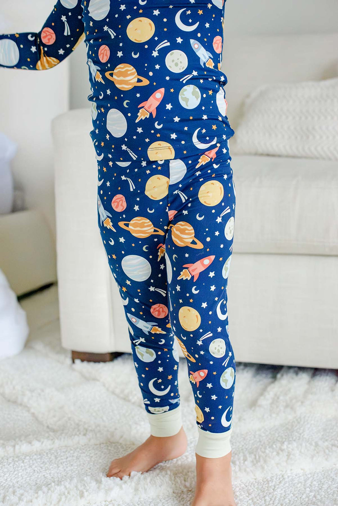 Close-up of a toddler’s legs on a shaggy white rug, wearing Caden Lane navy bamboo pajama pants with cream ankle cuffs in a Stellar Snuggles print featuring planets.