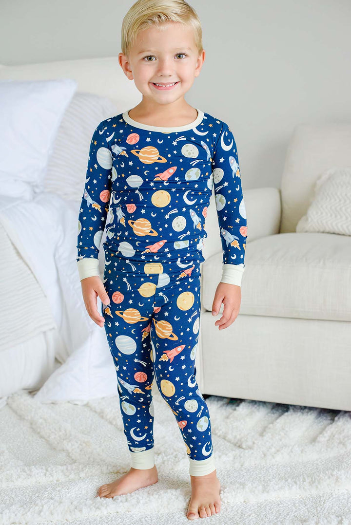 Full-length view of a smiling toddler standing on a shaggy rug by an armchair, wearing a Caden Lane navy bamboo pajama set in a Stellar Snuggles print featuring planets.