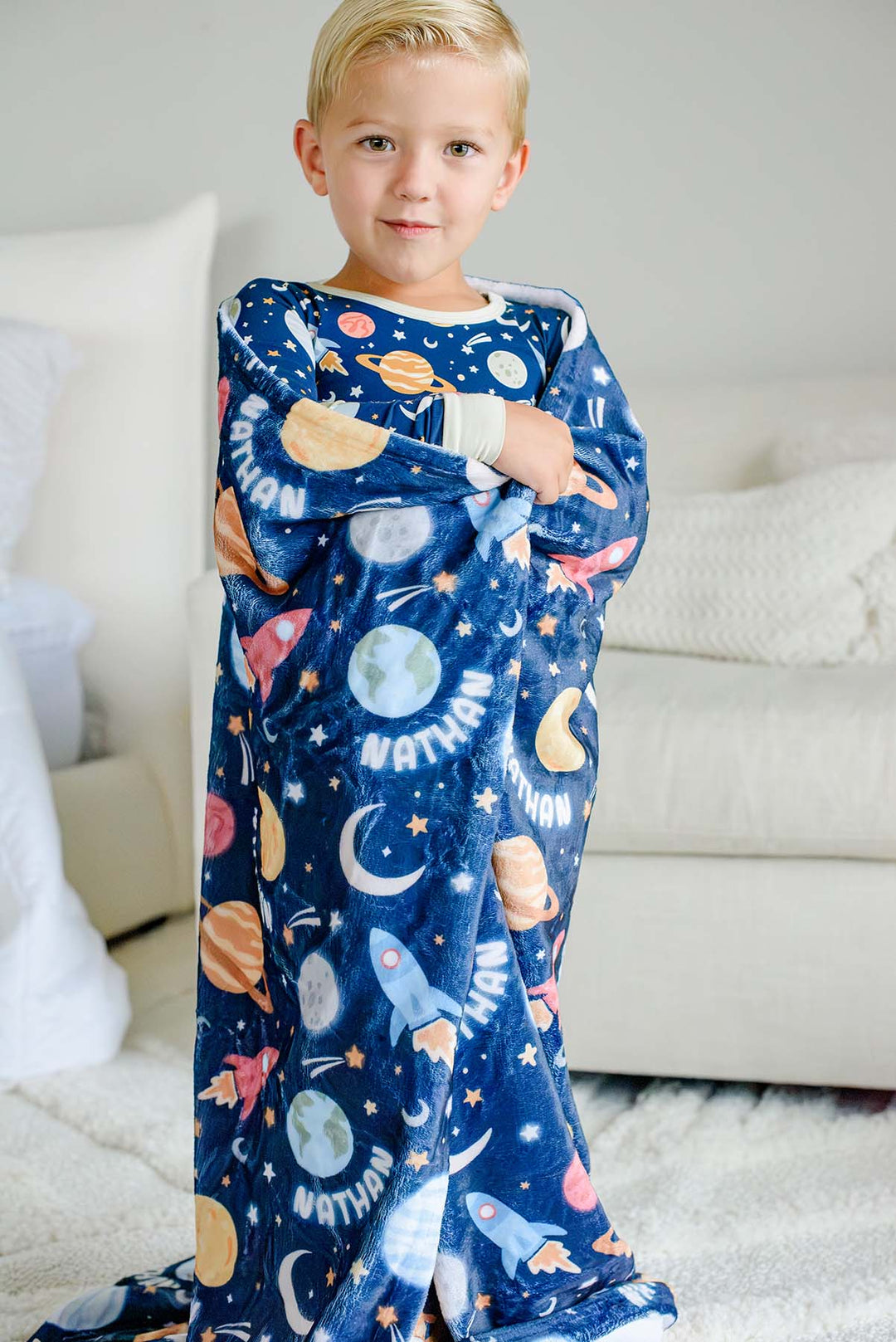 Full-length shot of a boy facing forward, wrapped in a Caden Lane navy plush blanket in a Stellar Snuggles print featuring moons and rockets, reading “Nathan,” paired with matching pajamas.