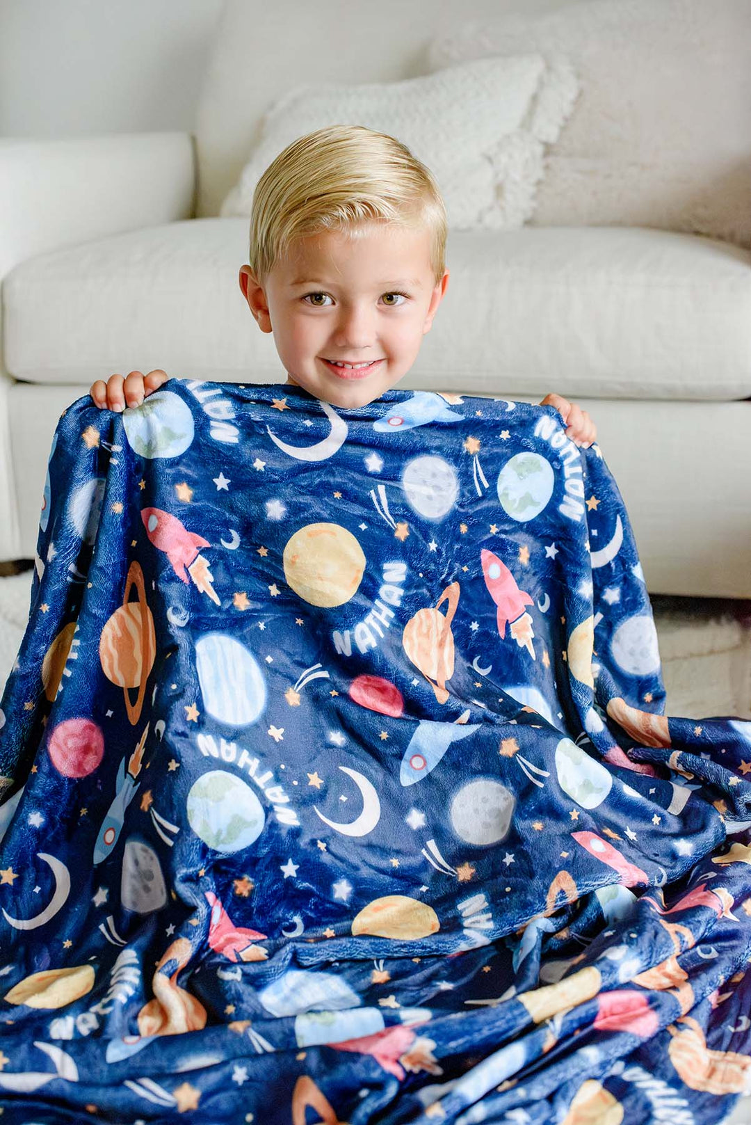 Grinning boy seated on the floor, holding up a Caden Lane navy plush blanket in a Stellar Snuggles print featuring planets, rockets, and stars, reading “Nathan.”