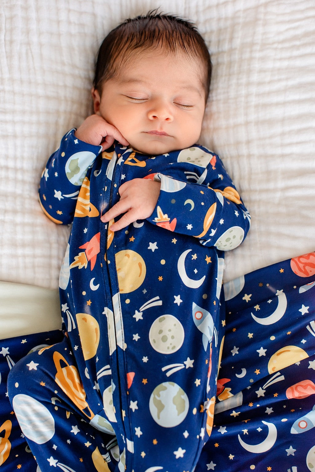 Overhead shot of an alert newborn cradled by adult hands, wearing a Caden Lane navy bamboo glow-in-the-dark zipper footie in a Stellar Snuggles print featuring planets and moons.
