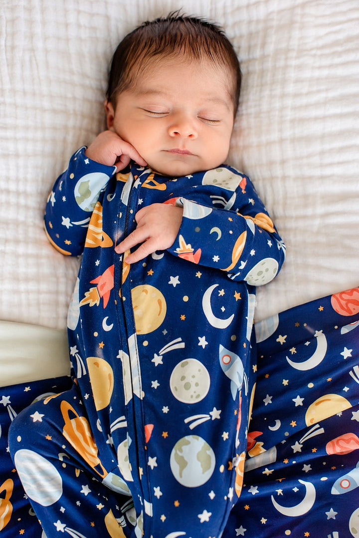 Overhead shot of an alert newborn cradled by adult hands, wearing a Caden Lane navy bamboo glow-in-the-dark zipper footie in a Stellar Snuggles print featuring planets and moons.