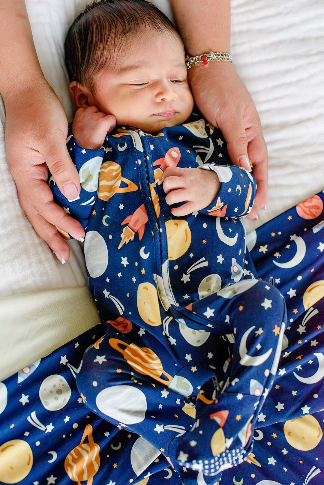 Overhead shot of a sleeping newborn with knees tucked up, wearing a Caden Lane navy bamboo glow-in-the-dark zipper footie in a Stellar Snuggles print featuring rockets and stars.