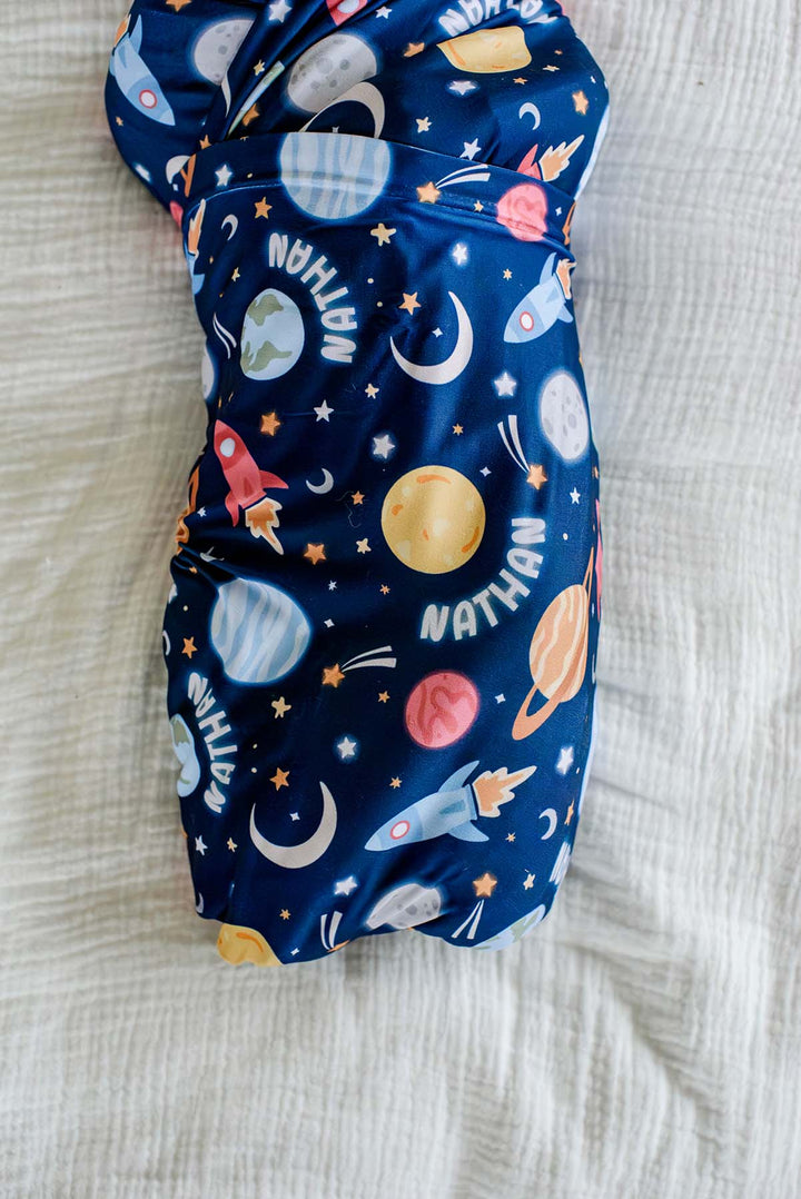 Close-up of a newborn, face cropped, wearing a Caden Lane navy knit swaddle in a Stellar Snuggles print featuring planets and rockets, reading “Nathan.”