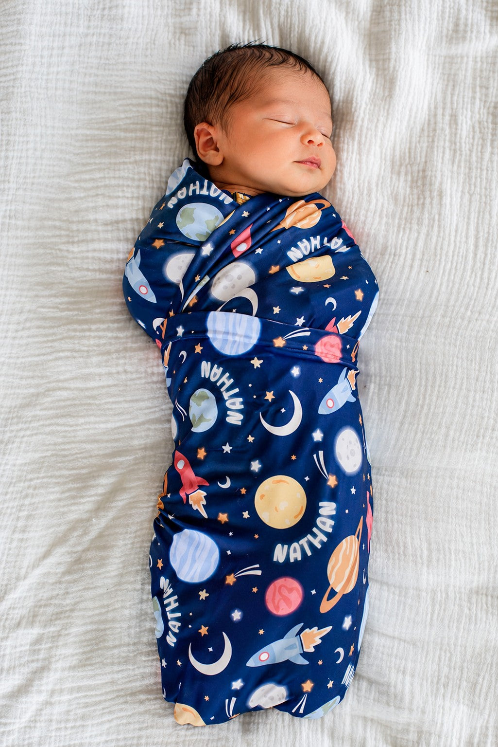 Full-length view of an alert newborn gazing upward, wearing a Caden Lane navy knit swaddle in a Stellar Snuggles print featuring moons, Saturn, and rockets, reading “Nathan.”