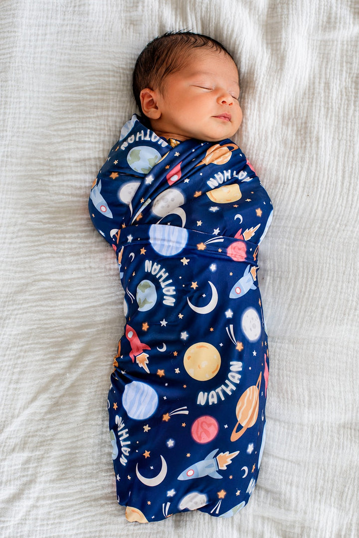 Full-length view of an alert newborn gazing upward, wearing a Caden Lane navy knit swaddle in a Stellar Snuggles print featuring moons, Saturn, and rockets, reading “Nathan.”