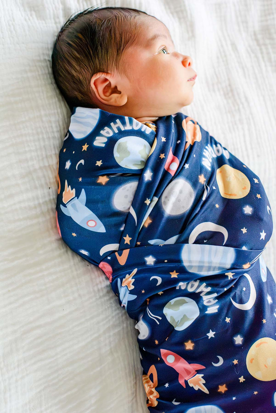 Side profile of an awake newborn wearing a Caden Lane navy knit swaddle in a Stellar Snuggles print featuring Earth, crescent moons, and rockets, reading “Nathan.”