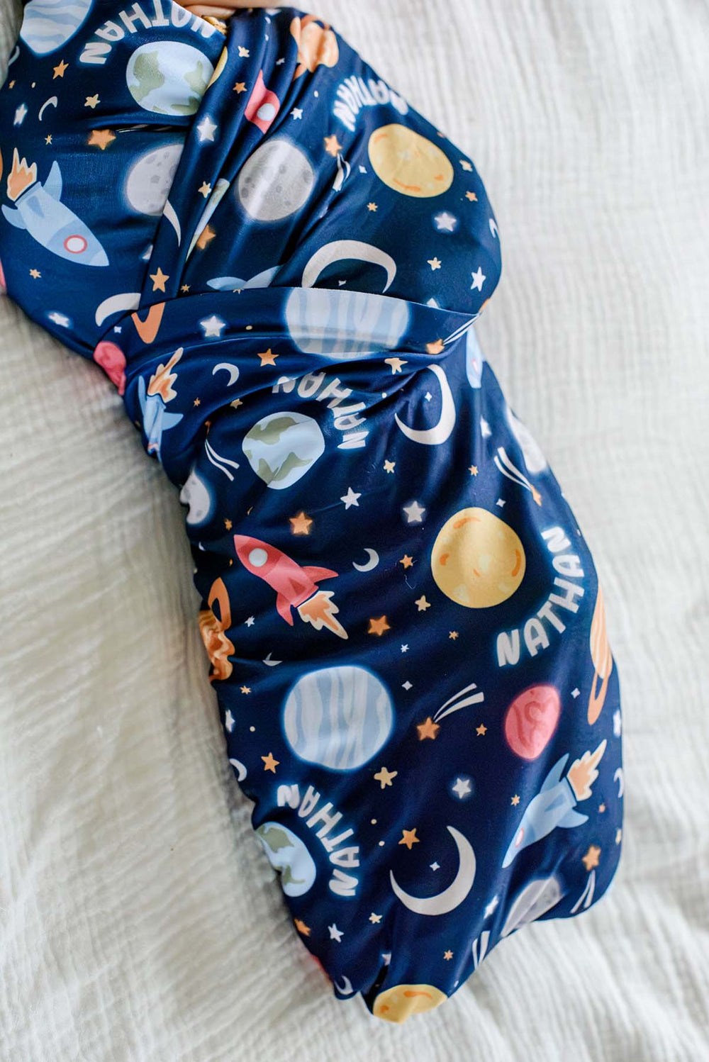Close-up of the wrapped folds on a newborn wearing a Caden Lane navy knit swaddle in a Stellar Snuggles print featuring planets and shooting stars, reading “Nathan.”