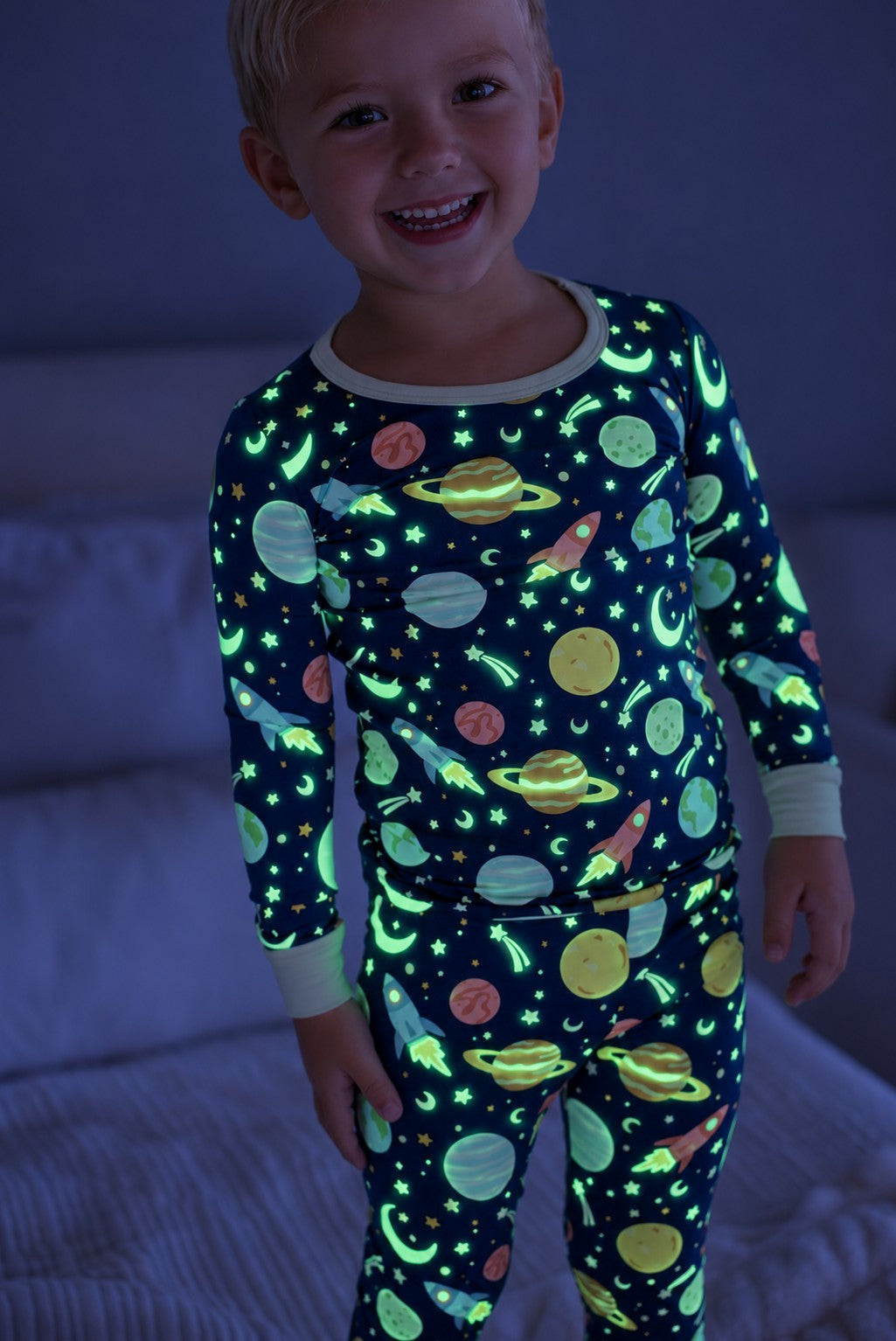 Smiling blond toddler standing by a bed, wearing a glowing Caden Lane navy bamboo two-piece pajama set in a Stellar Snuggles print featuring planets, rockets, and stars.