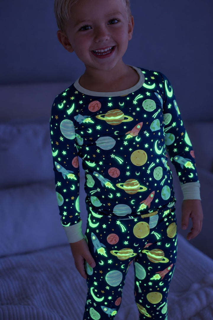 Smiling blond toddler standing by a bed, wearing a glowing Caden Lane navy bamboo two-piece pajama set in a Stellar Snuggles print featuring planets, rockets, and stars.