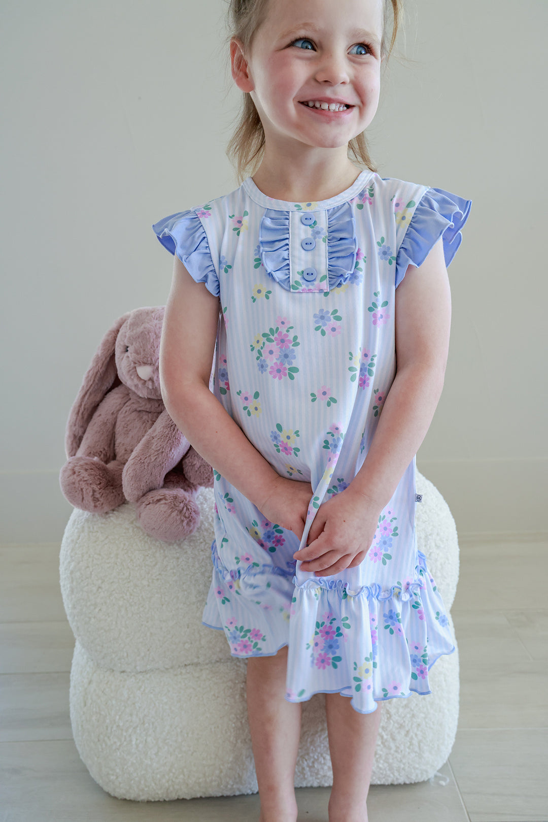 Young girl sitting on stool with stuffed bunny wearing Flutter Sleeve Nightgown in Heirloom Posies blue and white floral print, smiling full length view