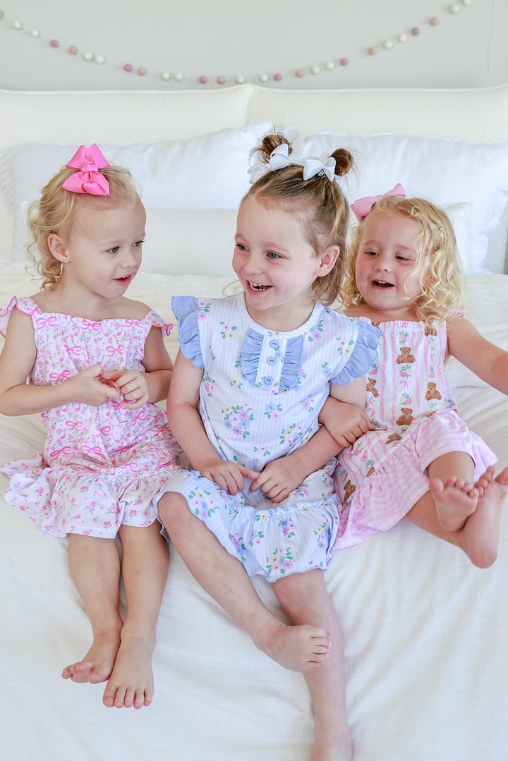 Three girls sitting on bed wearing Caden Lane summer nightgowns: Dreamy Bows pink bow print (left), Heirloom Posies blue floral (center), Beary Lovely teddy bear (right)