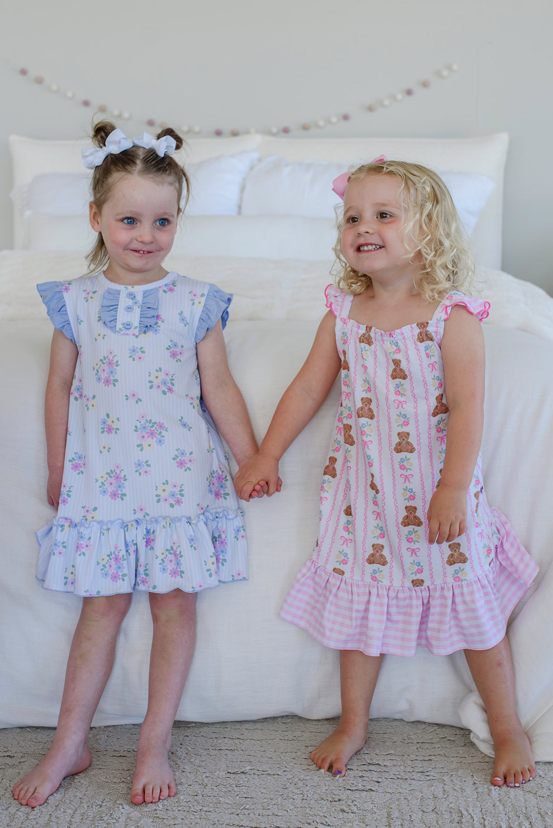 Two girls holding hands wearing summer nightgowns: Flutter Sleeve Nightgown in Heirloom Posies blue floral (left) and Beary Lovely teddy bear (right)