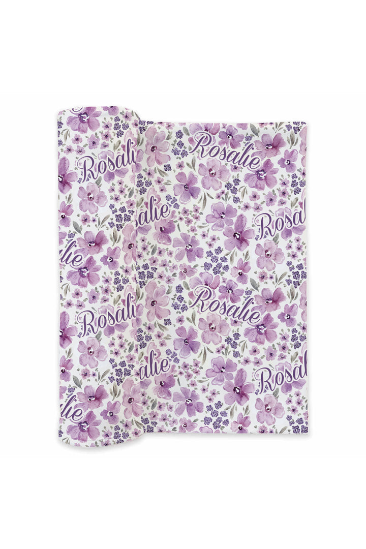 Flat lay of a partially unrolled Caden Lane white knit swaddle in a Lilac Dreams print featuring purple watercolor flowers and grey leaves, reading "Rosalie."