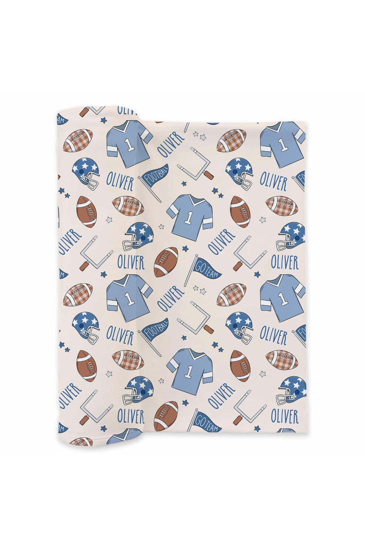 Caden Lane personalized swaddle blanket in blue football print, laid flat, showing jerseys, helmets, footballs, goal posts, and the custom name "Oliver" repeated throughout.