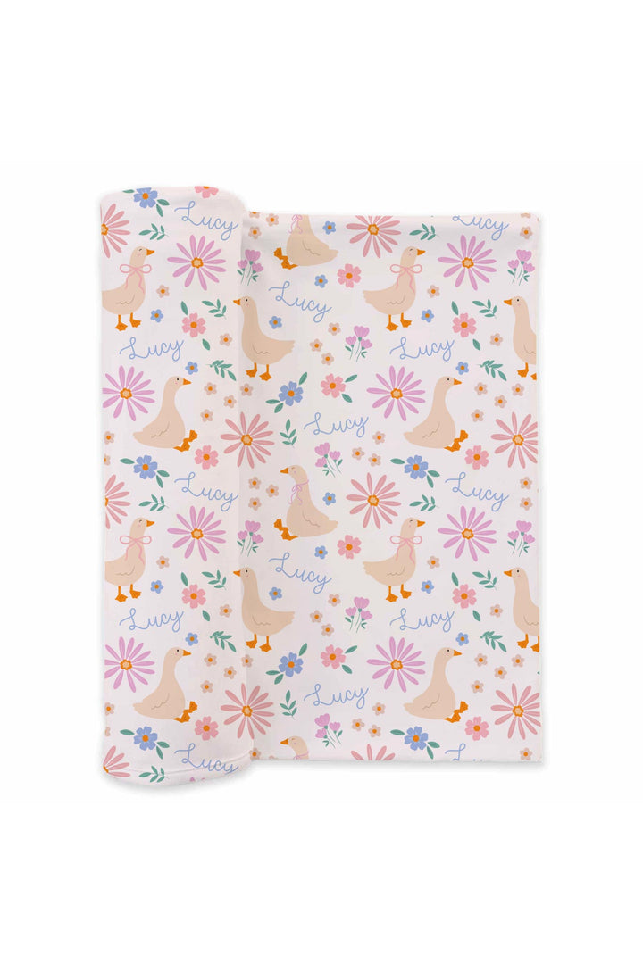Personalized Swaddle Blanket in the Lucy Goosy duck and floral print laid flat, showing geese, flowers, and name Lucy printed throughout on a white background