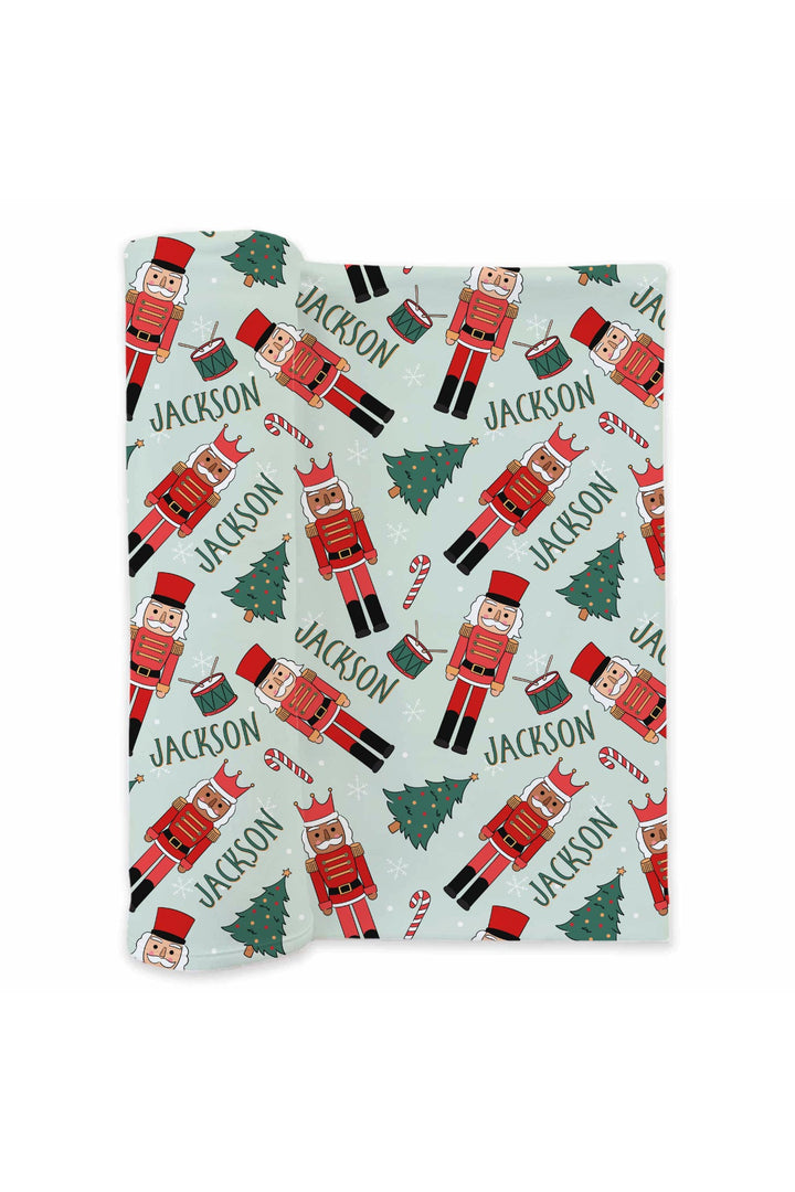Personalized Caden Lane Toy Soldier Green baby swaddle blanket featuring red nutcrackers, Christmas trees, candy canes, drums, and a custom name.