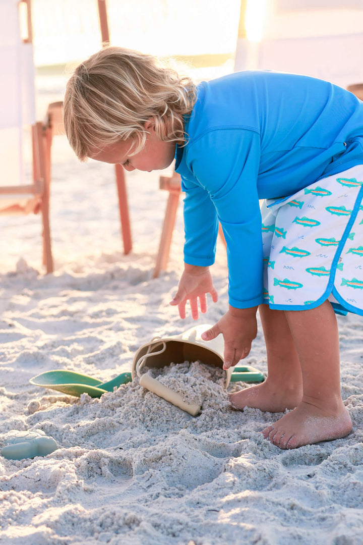 boy playing in sand in trout swim trunks and bright blue rash guard shirt 