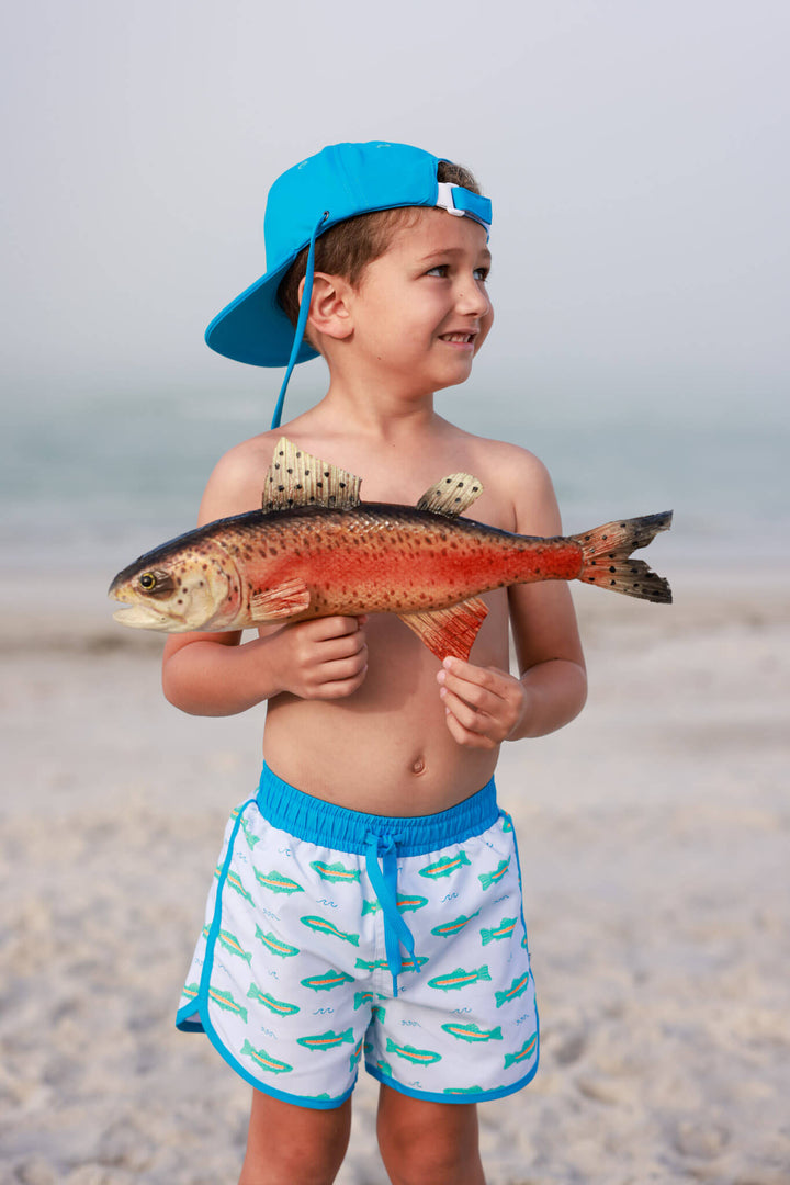 boy standing on beach holding a fish in tiny trout swim trunks with a matching blue hat 