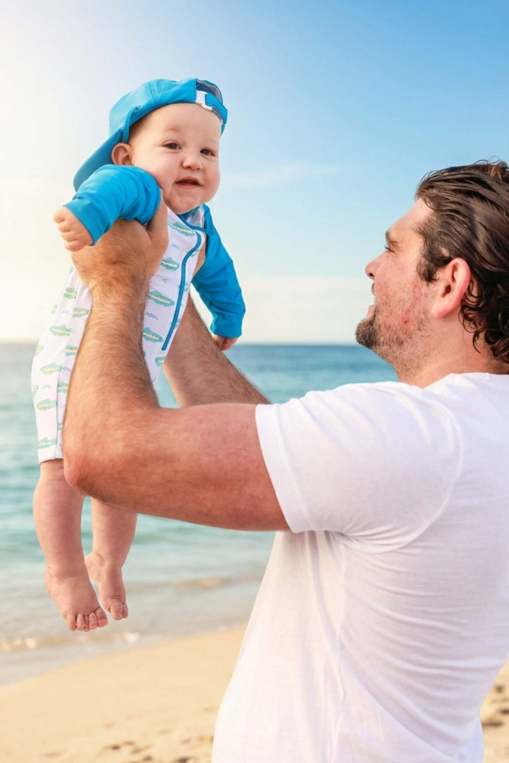 dad holding up baby on the beach wearing a trout long sleeve swim romper and matching surf hat 