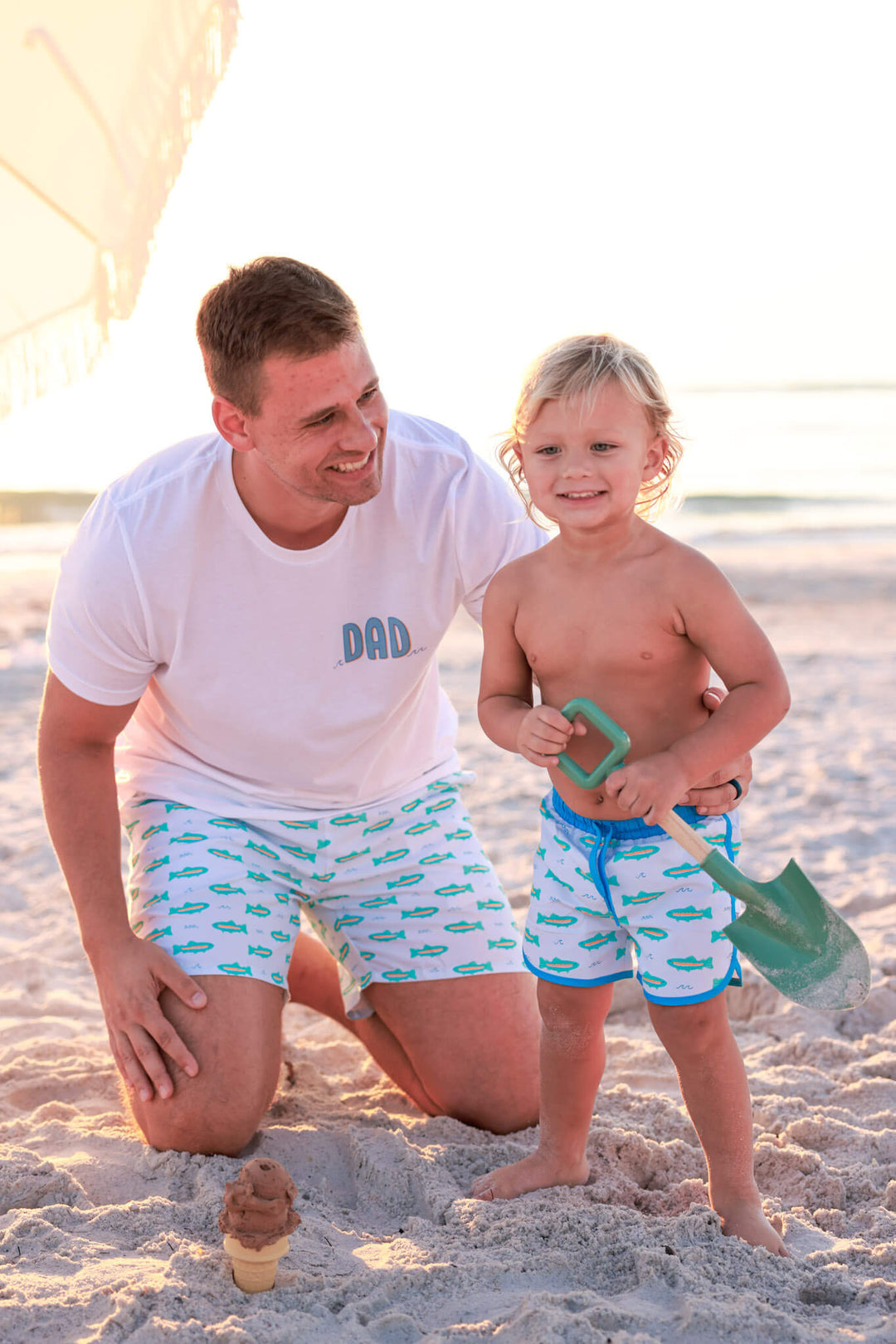 dad kneeling on beach with son in trout swim trunks with son in matching trunks holding a beach shovel 