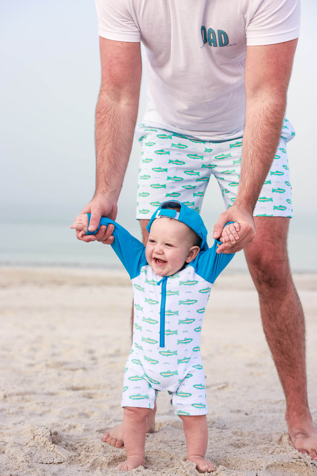dad helping baby walk on beach wearing matching tiny trout swimsuits 