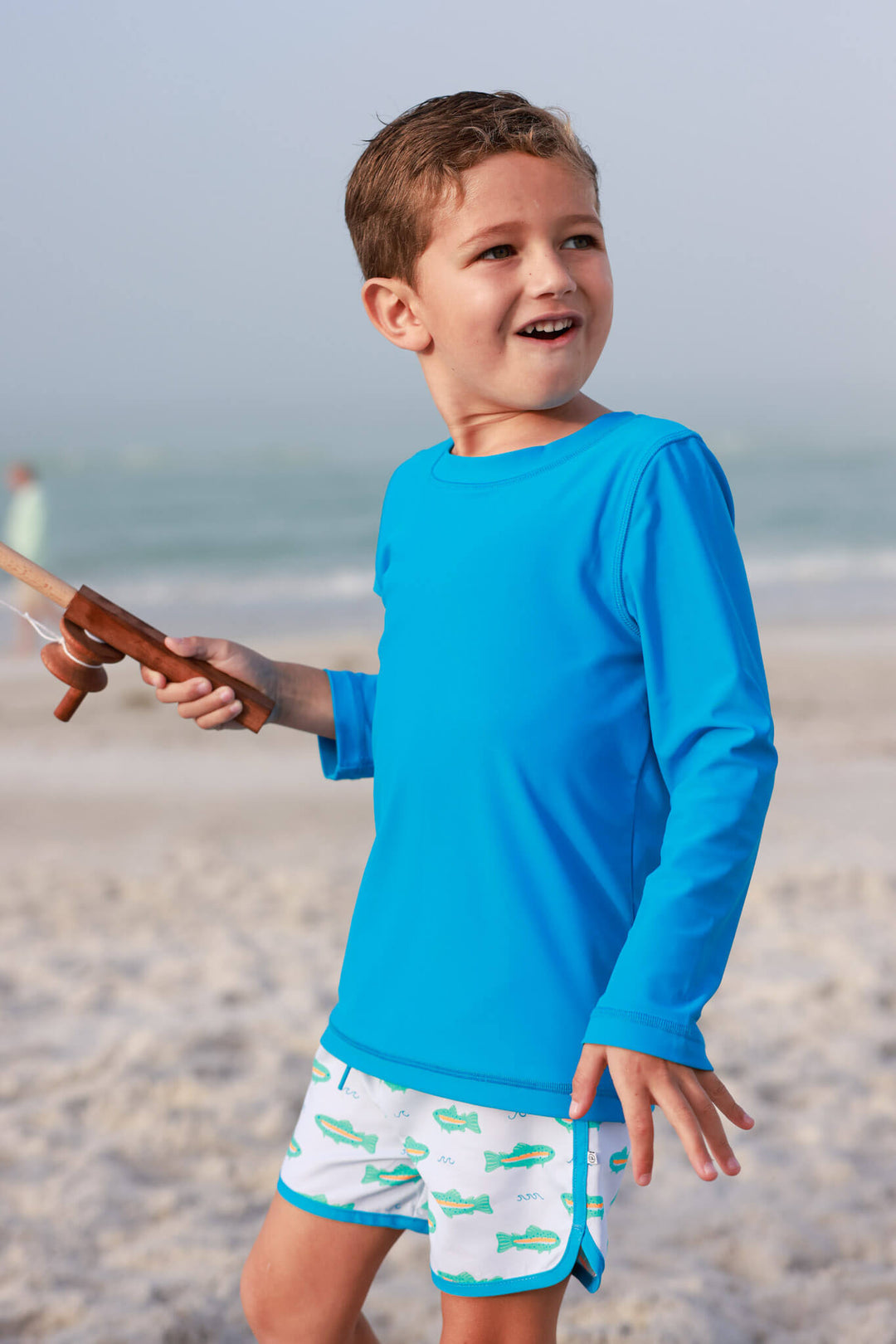 boy with fishing pole in hand in bright blue long sleeve rash guard shirt with trout swim trunks 