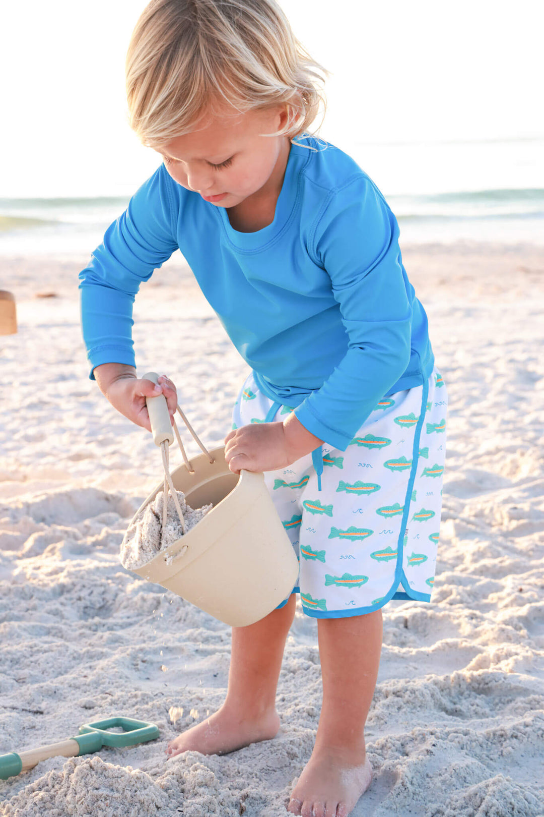 boy playing with beach bucket in twilight tide long sleeve rash guard shirt and tiny trout swim trunks 