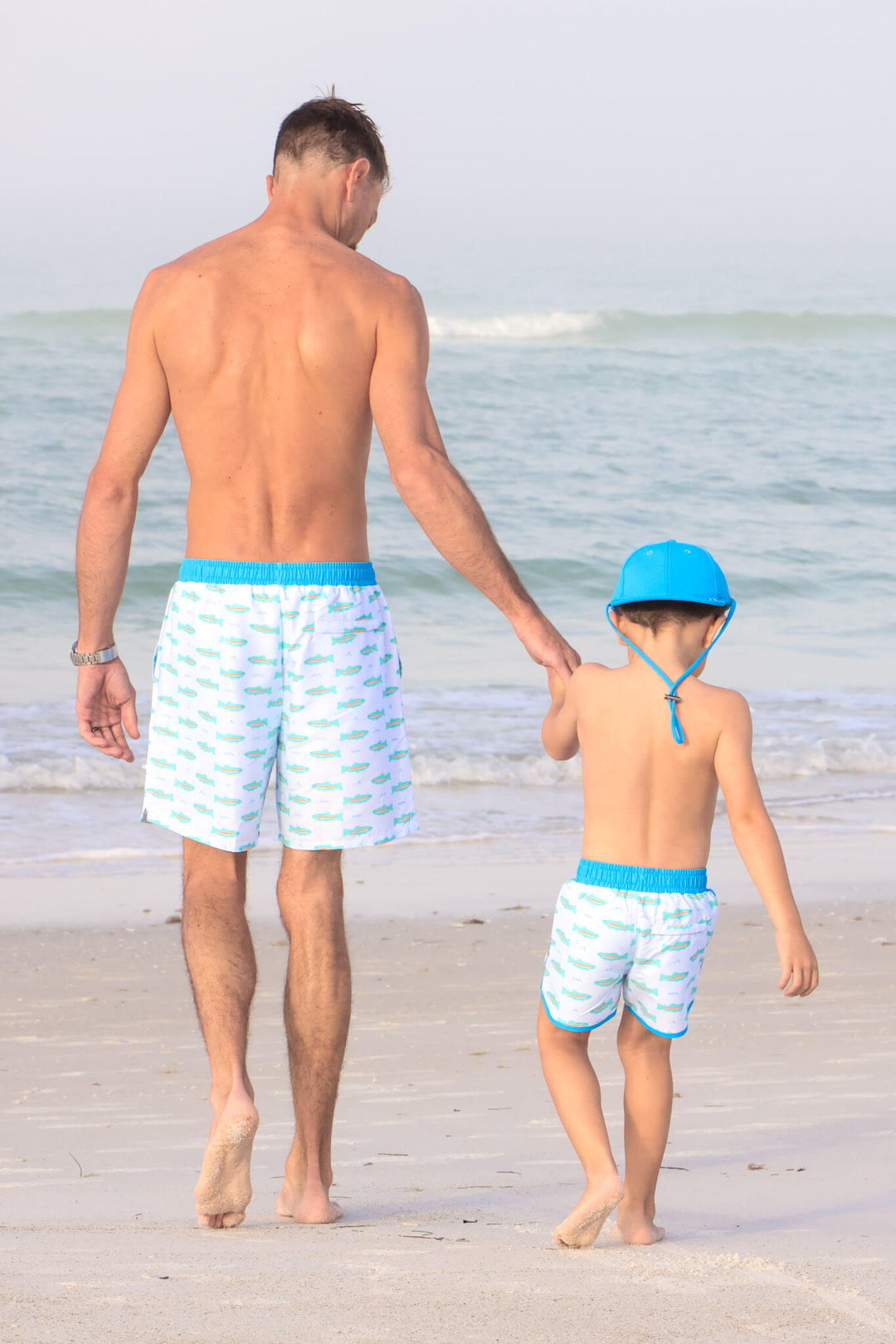 dad and son walking on beach in tiny trout swim trunks with a matching surf hat 