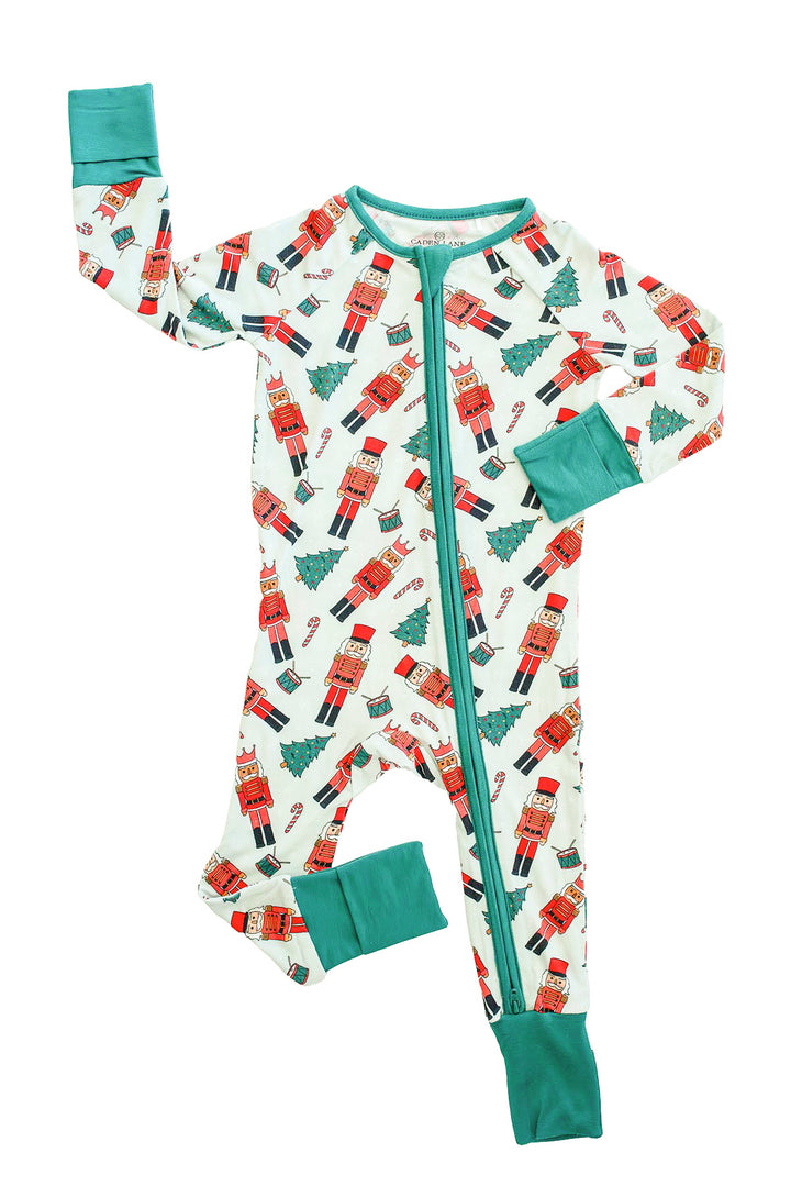 Flat lay of Caden Lane's white toy soldier print zip-up pajamas, featuring nutcrackers and Christmas trees, with green cuffs and trim.