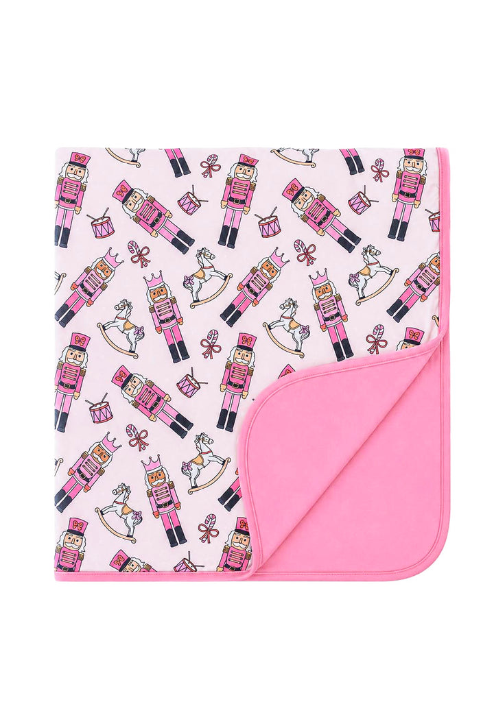 Caden Lane's pink toy soldier print baby blanket featuring nutcrackers, rocking horses, drums, and candy canes, with a solid pink reverse side, folded to show both sides.