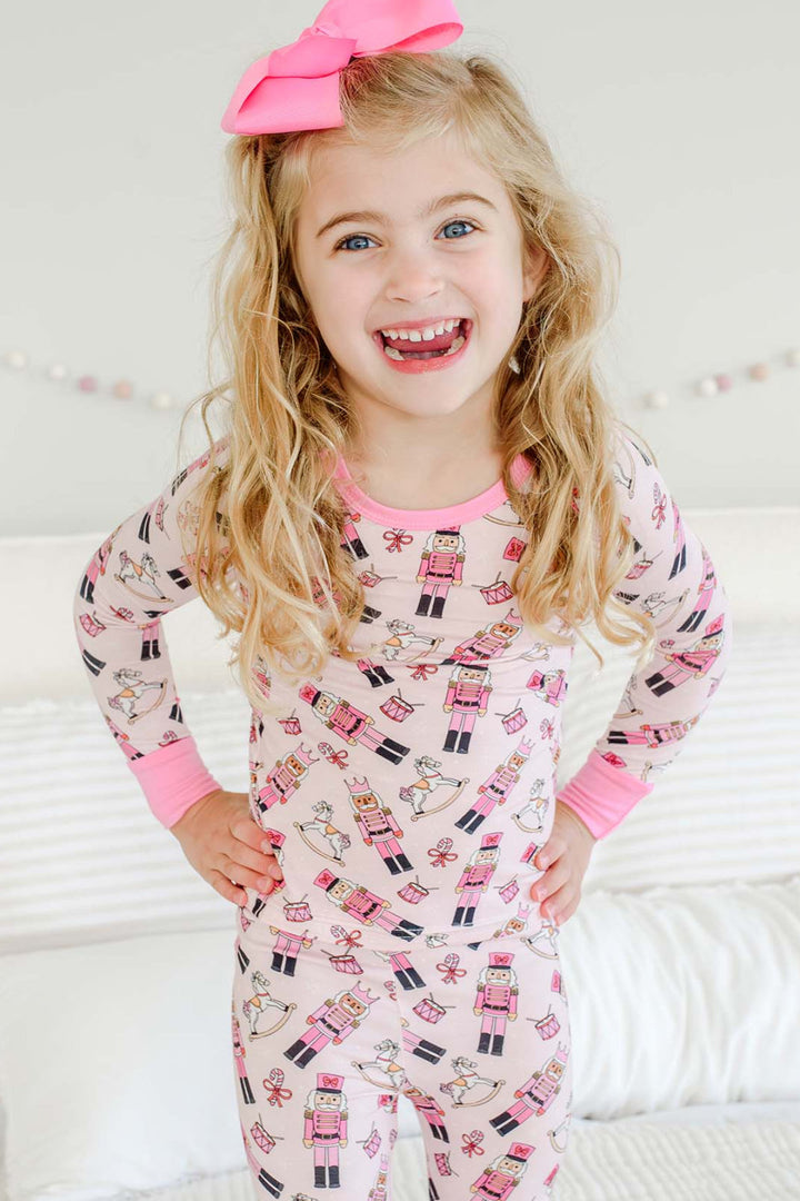 Girl smiling with hands on hips while wearing Caden Lane's pink toy soldier print pajamas, featuring nutcrackers, rocking horses, and candy canes, with a pink bow.