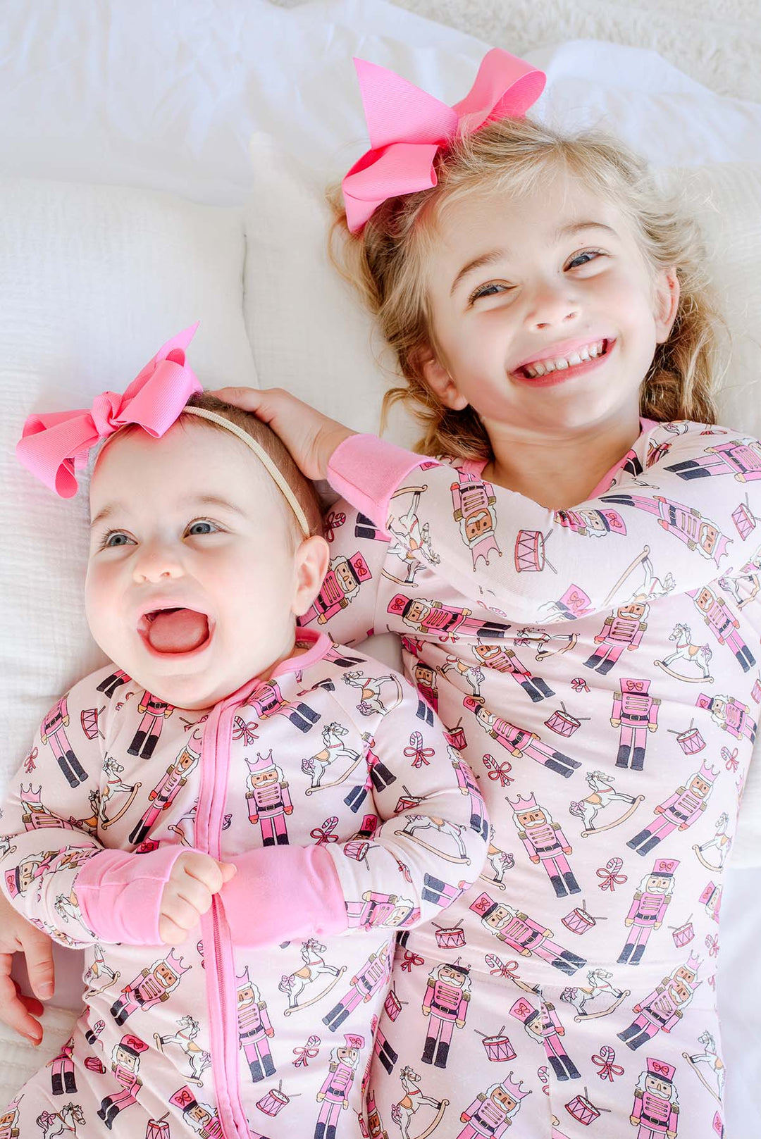 Two sisters wearing matching Caden Lane pink toy soldier print pajamas, featuring nutcrackers and rocking horses, with pink bows.
