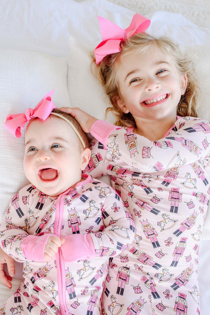 Two sisters wearing matching Caden Lane pink toy soldier print pajamas and rompers, featuring nutcrackers, rocking horses, and candy canes, with pink bows.