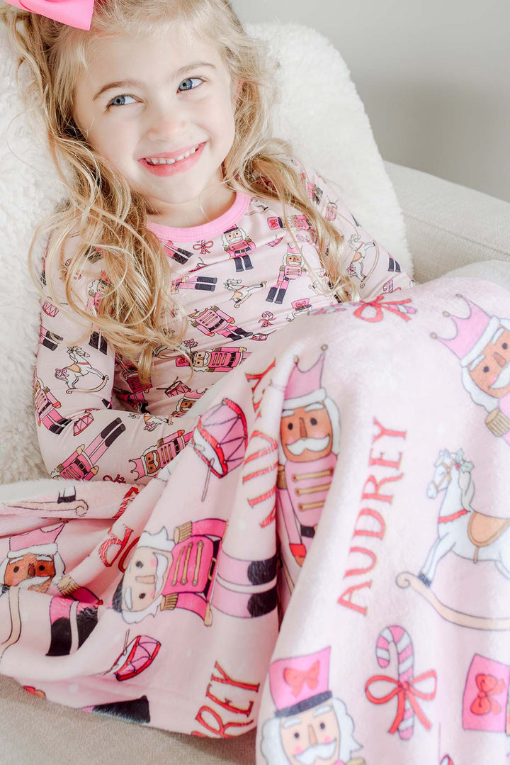 Girl wearing Caden Lane's pink toy soldier print pajamas, smiling while sitting with a matching personalized blanket featuring the name "Audrey."