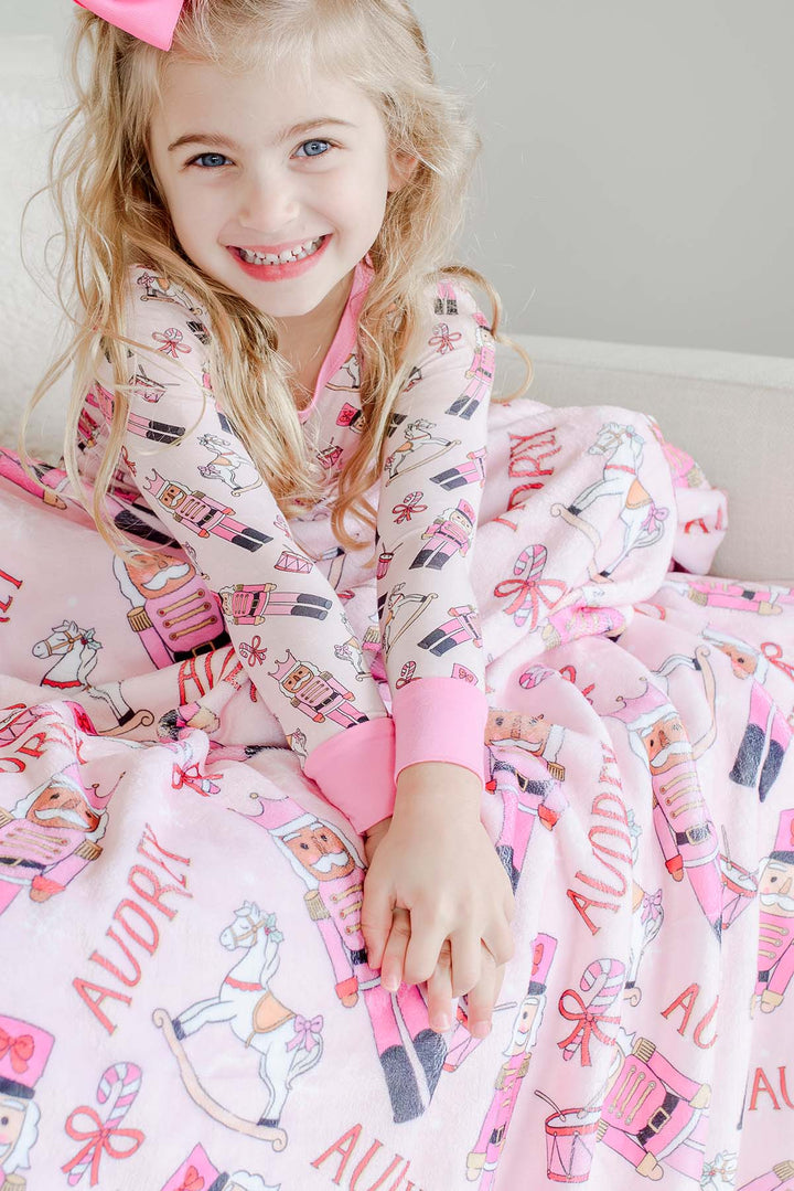 Girl wearing Caden Lane's pink toy soldier print pajamas, smiling while wrapped in a matching personalized blanket featuring the name "Audrey."