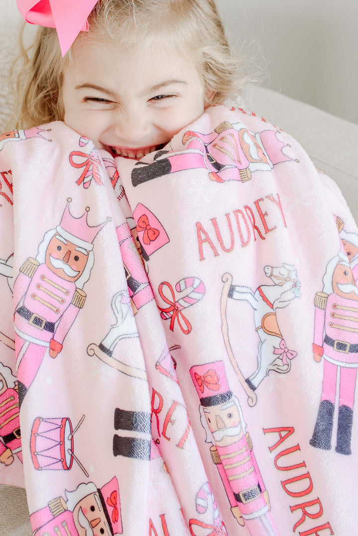 Close-up of a girl smiling while wrapped in Caden Lane's pink toy soldier print blanket personalized with the name "Audrey."