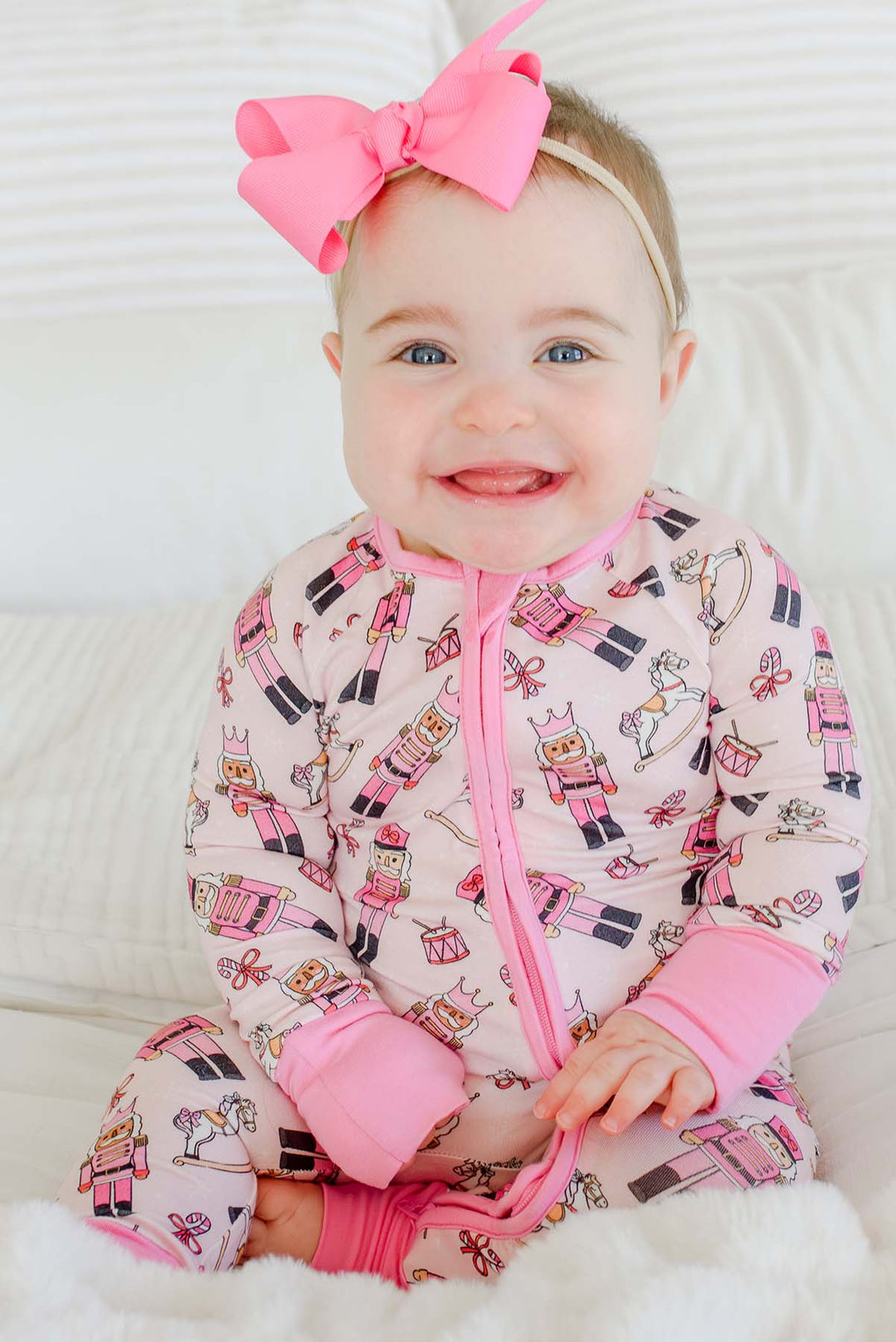 Baby girl smiling while wearing Caden Lane's pink toy soldier print zip-up pajamas, featuring nutcrackers and rocking horses, with a pink bow.