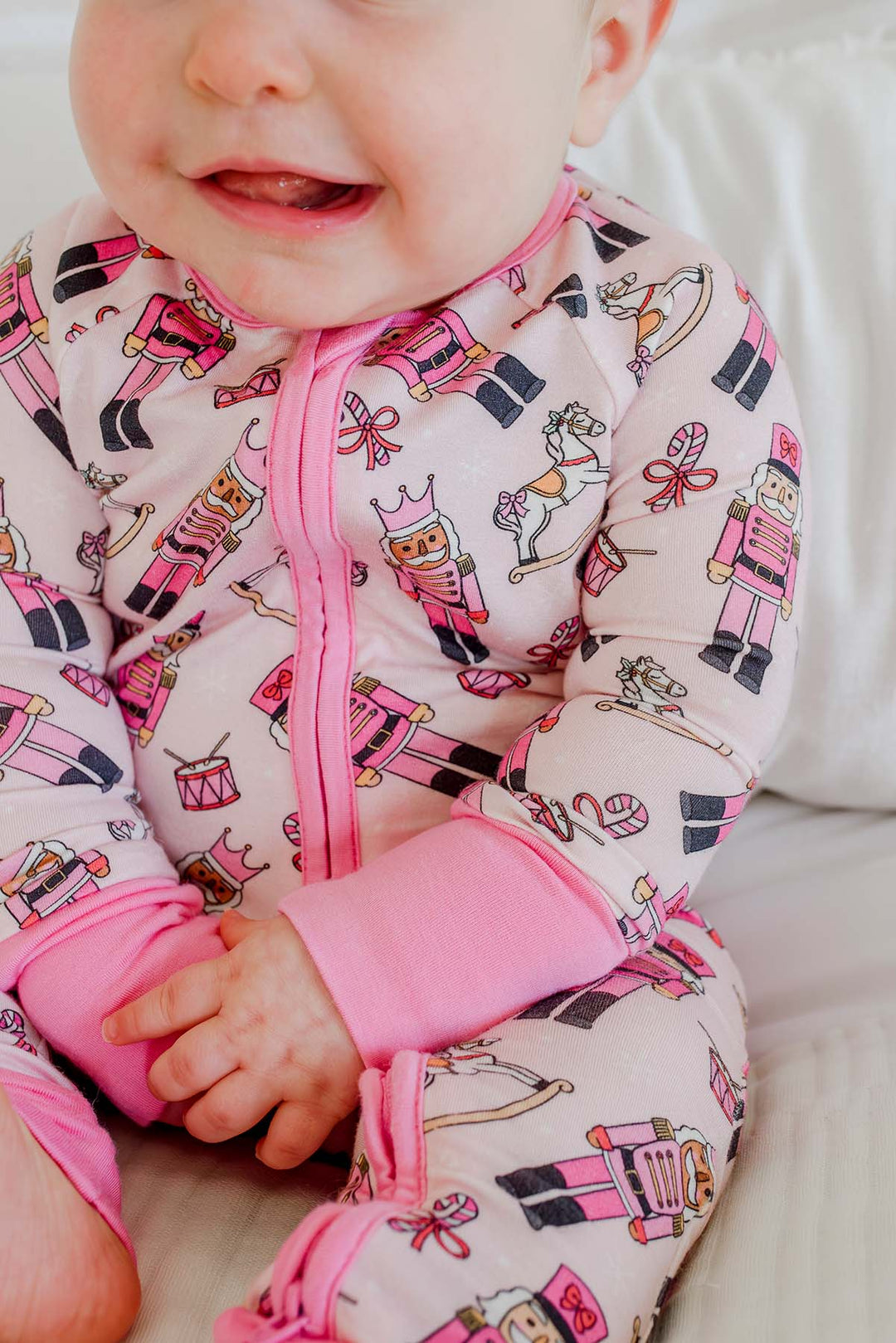 Close-up of baby girl wearing Caden Lane's pink toy soldier print zip-up pajamas, featuring nutcrackers and rocking horses, with pink cuffs.