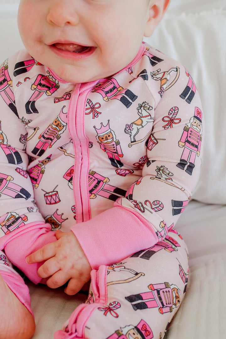 Close-up of baby girl wearing Caden Lane's pink toy soldier print zip-up pajamas, featuring nutcrackers and rocking horses, with pink cuffs.