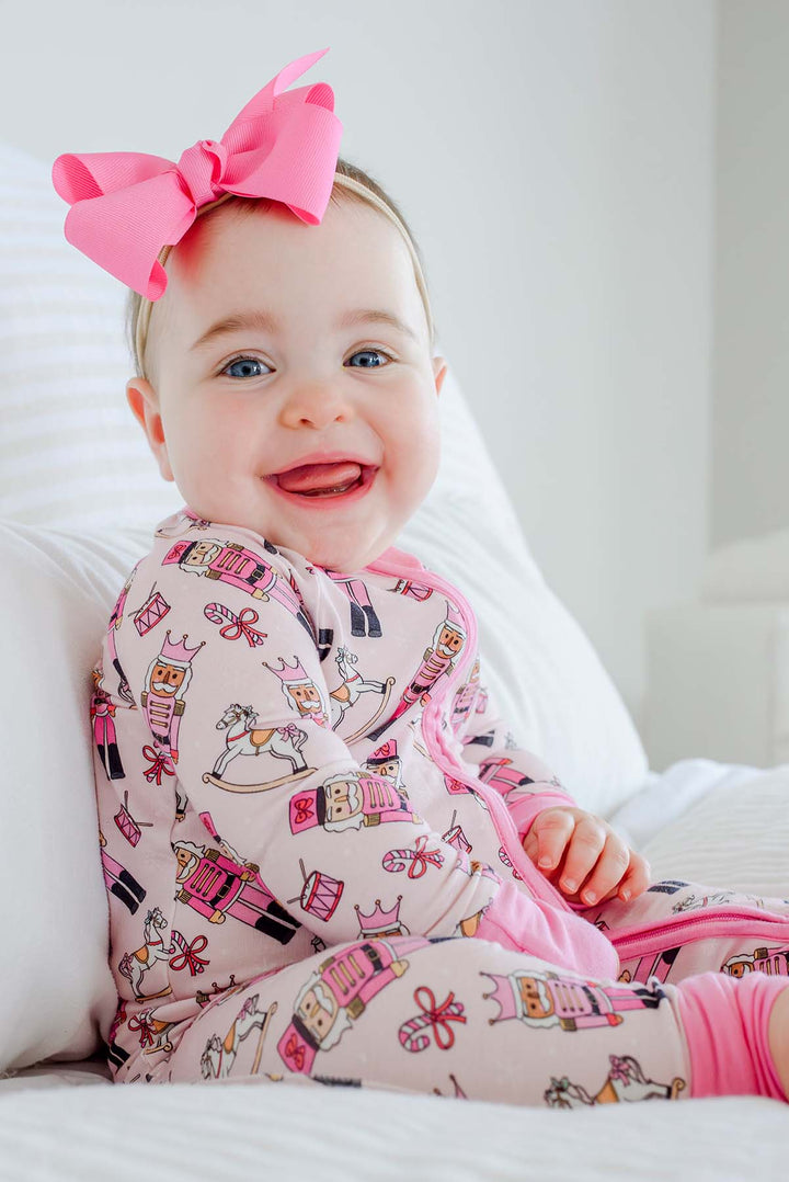 Baby girl smiling while wearing Caden Lane's pink toy soldier print zip-up pajamas, featuring nutcrackers and rocking horses, with a pink bow.