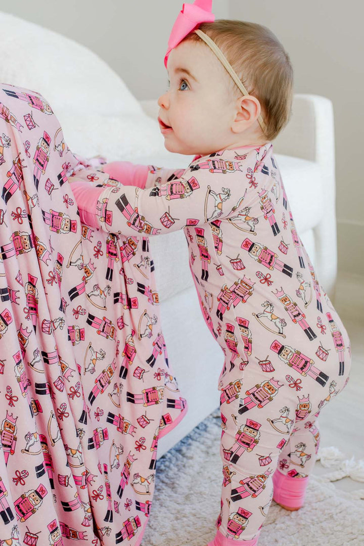 Baby girl standing while wearing Caden Lane's pink toy soldier print zip-up pajamas, featuring nutcrackers and rocking horses, with a pink bow.