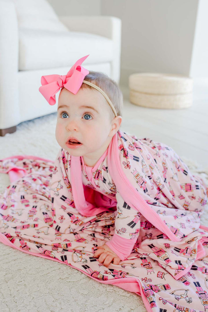 Baby wearing a pink bow and Caden Lane's pink toy soldier print zip-up sleeper, crawling on a matching blanket featuring nutcrackers, rocking horses, and candy canes.