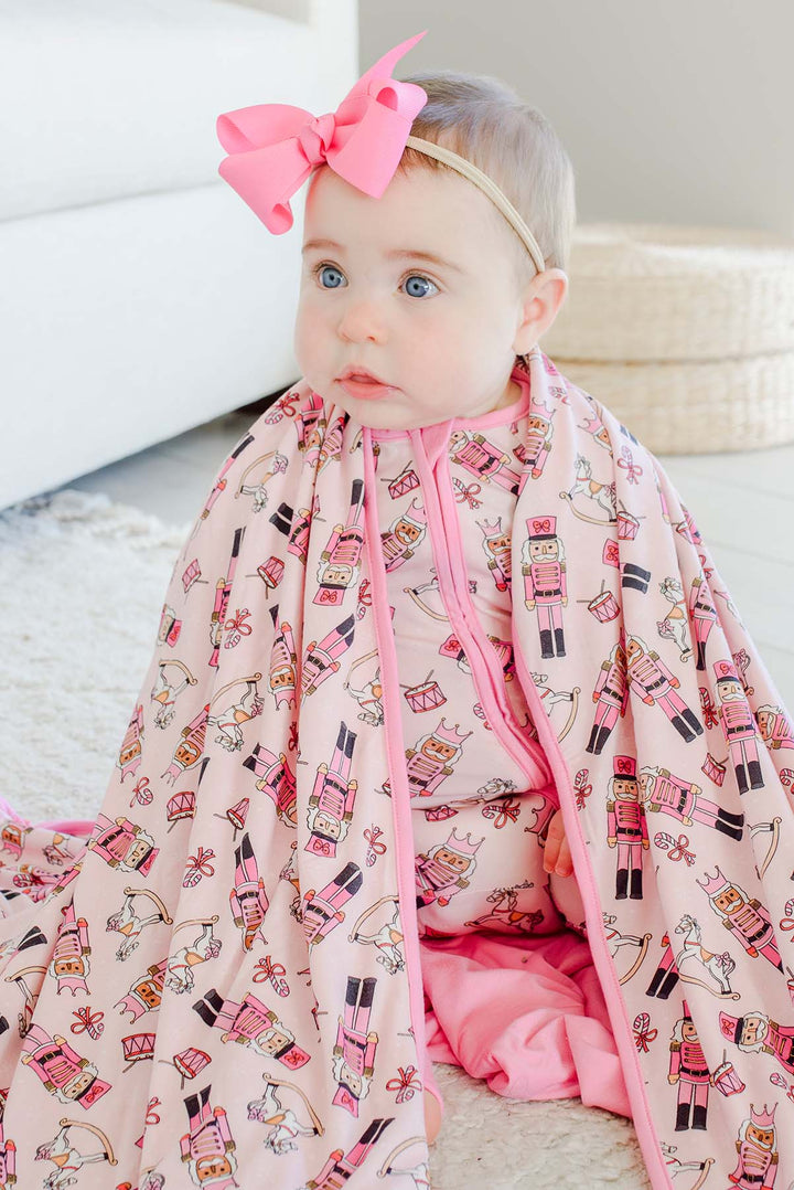 Baby wearing a pink bow and Caden Lane's pink toy soldier print zip-up sleeper, wrapped in a matching blanket featuring nutcrackers, rocking horses, and candy canes.