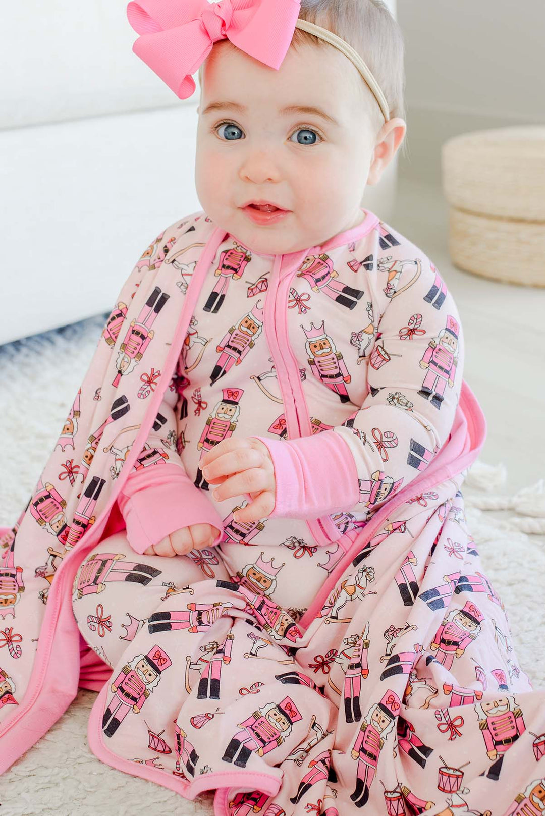 Close-up of a baby wearing a pink bow and Caden Lane's pink toy soldier print zip-up sleeper, wrapped in a matching blanket featuring nutcrackers, rocking horses, and candy canes.