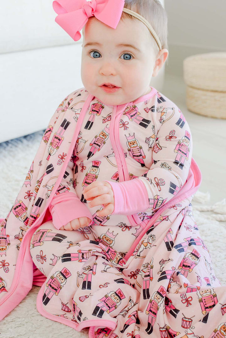Close-up of a baby wearing a pink bow and Caden Lane's pink toy soldier print zip-up sleeper, wrapped in a matching blanket featuring nutcrackers, rocking horses, and candy canes.
