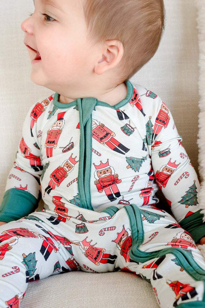 Close-up of baby boy smiling while wearing Caden Lane's white toy soldier print zip-up pajamas, featuring nutcrackers and Christmas trees, with green cuffs.