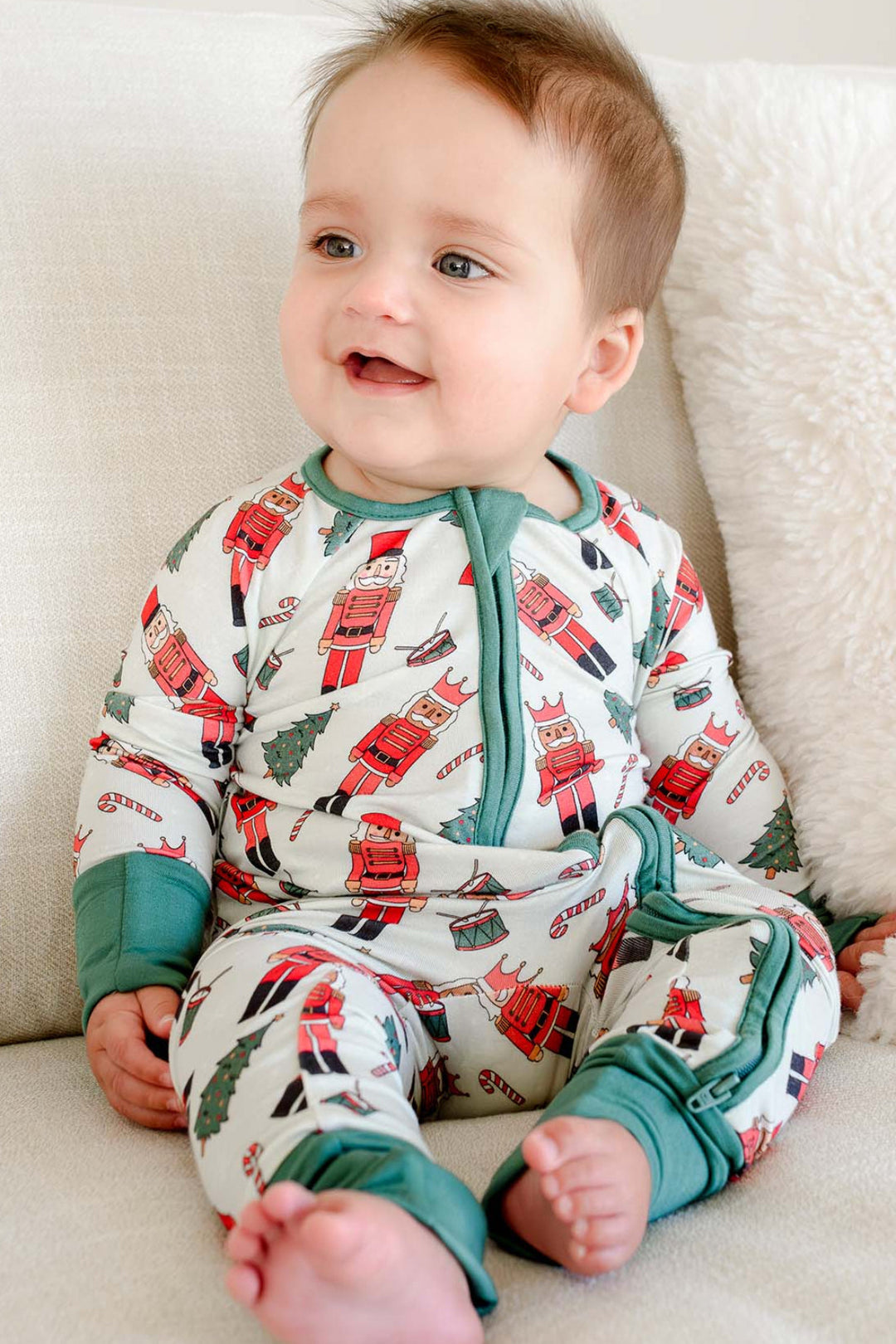 Baby boy sitting while wearing Caden Lane's white toy soldier print zip-up pajamas, featuring nutcrackers and Christmas trees, with green cuffs.