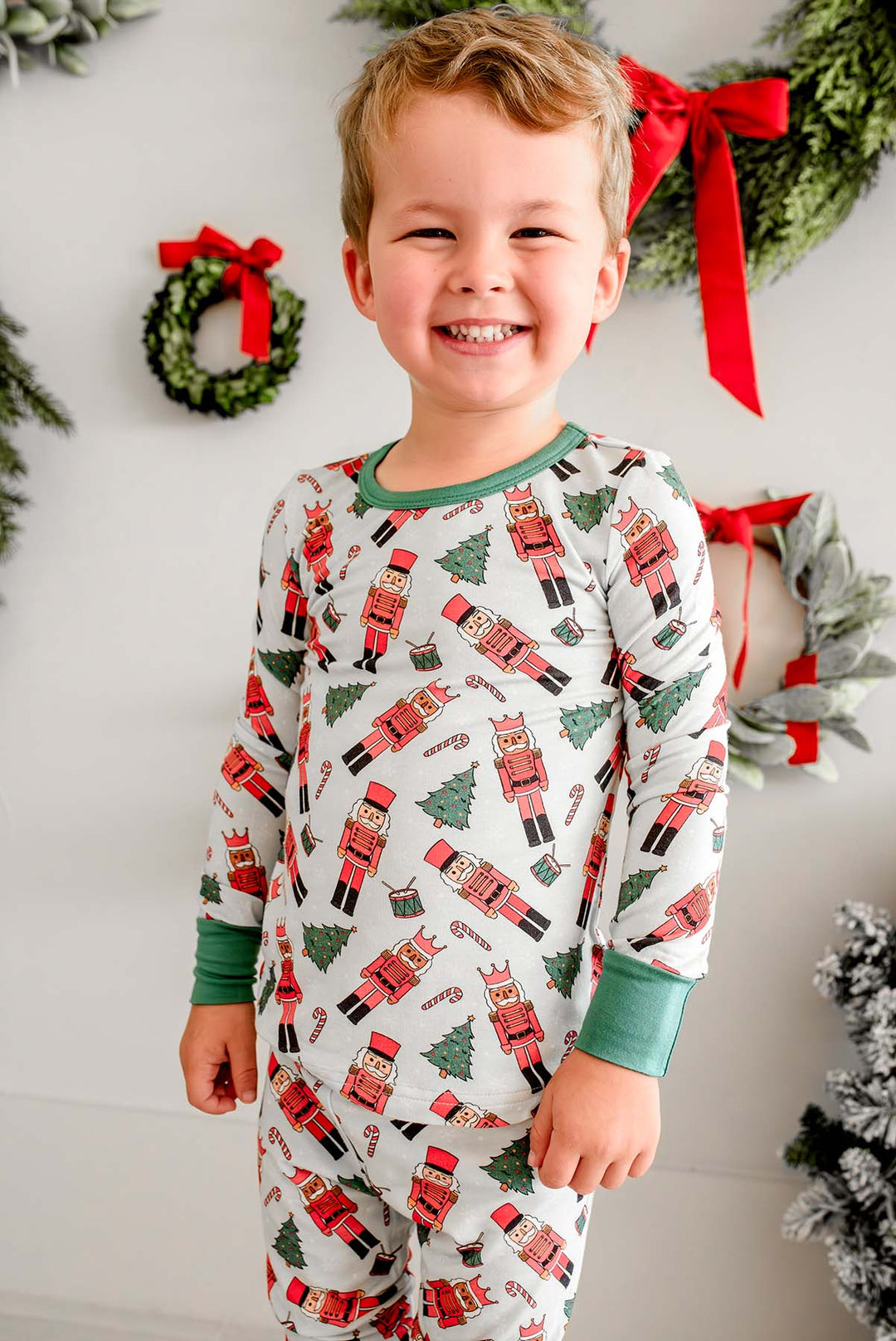Boy smiling while wearing Caden Lane's white toy soldier print pajamas, featuring nutcrackers and Christmas trees, with green cuffs, standing in front of wreaths.