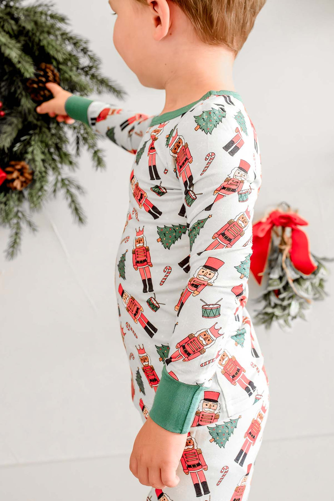 Close-up of boy wearing Caden Lane's white toy soldier print pajamas, featuring nutcrackers and Christmas trees, with green cuffs, reaching toward a wreath.